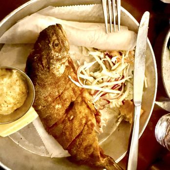 Work & Class - Whole Fried Idaho Trout - The image shows a whole fried Idaho trout placed on a metal plate lined with paper. The plate also contains a serving of coleslaw, a bread roll, and a small metal container of dipping sauce. A fork and knife are positioned on the plate, ready for eating. The setting appears to be a casual dining environment. - New American Near Me - Denver, Colorado