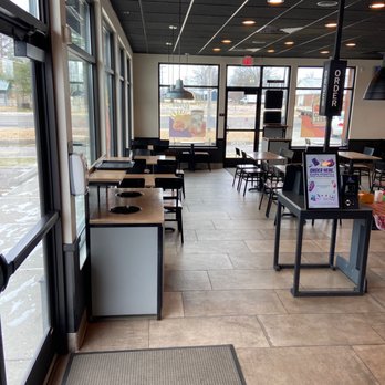 Taco Bell - Lobby entrance. - The photo depicts the interior of a Taco Bell restaurant featuring a seating area with multiple tables and chairs. Large windows on the left side allow natural light to illuminate the space, and a menu stand is positioned near the entrance. The ceiling has a grid pattern with recessed lighting, and the floor is tiled with large, rectangular tiles. The overall setting appears clean and modern, with a view of the outside street through the windows. - Tacos Near Me - Cuba, Missouri
