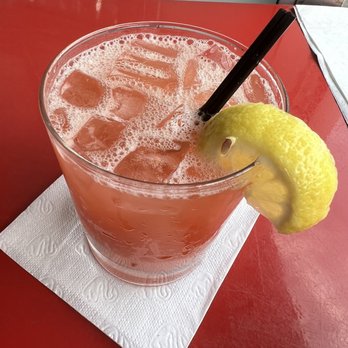 Sugarcane - Sugarcane Frozen Rum Punch - A clear glass contains a frozen rum punch with ice cubes and foam on top. The drink is garnished with a lemon wedge on the rim and has two black straws inserted. It is set on a white paper napkin on a red table, with a background that includes a partially visible wall and some papers. - Caribbean Near Me - Brooklyn, New York
