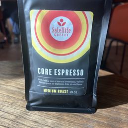 Core Espresso Beans were delicious! - A black resealable bag of Satellite Coffee Core Espresso beans, labeled as medium roast, sitting on a wooden table in a coffee shop setting.