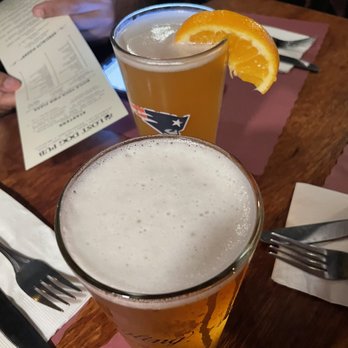 Lost Dog Pub - Wide selection of tasty beer options. - Two glasses of beer are placed on a wooden table at a restaurant, one with a foam head and the other garnished with an orange slice. - American Near Me - South Dennis, Massachusetts