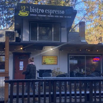 Bistro Espresso - Front entrance - The image shows the exterior of Bistro Espresso, a family-owned restaurant established in 1993, with outdoor seating on a wooden deck. A woman stands near the entrance, which has a sign for additional backyard seating. The storefront displays a neon 'Open' sign, and the building is surrounded by trees with autumn foliage. The signage indicates offerings such as espresso, restaurant meals, pastries, beer, wine, and ice cream. - Coffee & Tea Near Me - Arnold, California