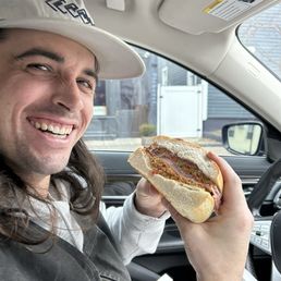 HALF of a large muffuletta - A man wearing a white cap and a black vest is sitting in a car, smiling, and holding a sandwich with meat and vegetables.