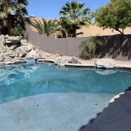 Aqua Shine ground down all the pebble that was above the surface and replaced it with flagstone that amazingly matches our current decking - A backyard swimming pool features rocks, palm trees, and a concrete deck, with a privacy wall and shadows cast by trees.