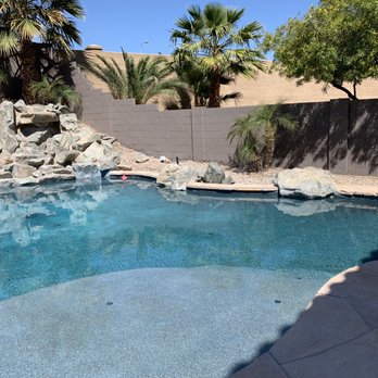 Aqua Shine Pool Services - Aqua Shine ground down all the pebble that was above the surface and replaced it with flagstone that amazingly matches our current decking - A backyard swimming pool features rocks, palm trees, and a concrete deck, with a privacy wall and shadows cast by trees. - Pool Cleaners Near Me - Phoenix, Arizona