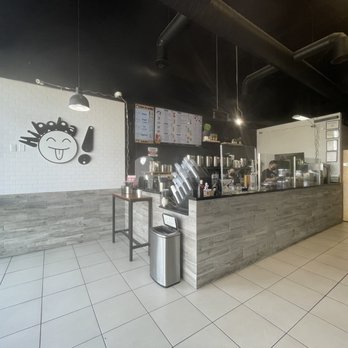 Hi BoBa - The image shows the interior of a bubble tea shop with a counter area where staff prepare drinks. The counter is covered with gray tiles and has various equipment and supplies. Behind the counter, there are shelves with containers and ingredients, and a menu display on the wall. To the left, a large window allows natural light into the space. The wall features a white tile section with a black cartoon face and an exclamation mark. The ceiling is black with visible ductwork and hanging lights, and the floor is tiled with large, light-colored tiles. - Bubble Tea Near Me - Albuquerque, New Mexico