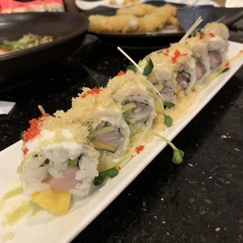 Miso - Sexy Lazy Roll with yellowtail - Sushi rolls with fish, vegetables, and toppings are arranged on a white rectangular plate, with other dishes and a child visible in the background. - Japanese Near Me - Round Rock, Texas
