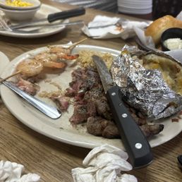 A restaurant plate holds a partially eaten steak, shrimp skewers, and a baked potato wrapped in foil, with utensils and crumpled napkins on a wooden table.