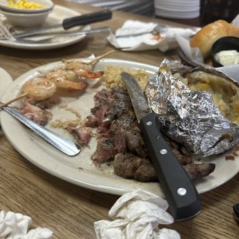 Logan's Roadhouse - A restaurant plate holds a partially eaten steak, shrimp skewers, and a baked potato wrapped in foil, with utensils and crumpled napkins on a wooden table. - Steakhouses Near Me - Flowood, Mississippi