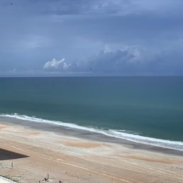 A photograph captures a coastal scene with a sandy beach, ocean waves, and a cloudy sky. The view is from an elevated position, showing the shoreline, the water, and the horizon. The sky is overcast with dark clouds, indicating possible rain or stormy weather. The beach appears mostly empty with minimal human activity visible.