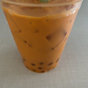 Tong Phoon - Thai Tea with Bubbles - The image shows a transparent plastic cup filled with Thai iced tea, which has an orange-brown color and contains ice cubes and black tapioca pearls at the bottom. A green straw is inserted into the drink, and the cup is placed on a light-colored surface. The background is blurred, indicating an indoor setting. - Thai Near Me - Bristol, Rhode Island