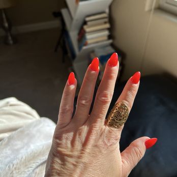 Flashy Nails - A hand with long, bright red painted nails is displayed, wearing a large gold ring with an intricate design, in front of a bookshelf filled with books. - Nail Salons Near Me - Albuquerque, New Mexico