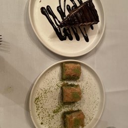 Pistachio Baklava - The image shows two white plates on a table with desserts. The top plate has a slice of chocolate cake decorated with chocolate drizzle on a white plate. The bottom plate contains three pieces of pistachio baklava, garnished with green pistachio powder. The table surface is light-colored, and a glass of water is partially visible in the lower right corner.