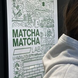 The sign features the name 'Matcha Matcha Lab' in bold green letters, with a detailed line drawing above and around it. The illustration includes trees, leaves, a teapot, and other nature-inspired elements, creating a thematic connection to matcha and natural ingredients. The sign appears to be indoors, with a person partially visible in front of it, wearing a light-colored hoodie.