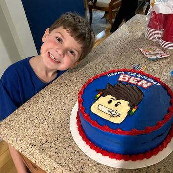 Benny's Bakery - C - A young boy with short brown hair and a wide smile stands beside a birthday cake on a kitchen counter. The cake is blue with red piping and features a Lego character face with the name 'Ben' and the message 'Happy 7th Birthday'. In the background, there are chairs, a table, and party supplies, indicating a birthday celebration. - Bakeries Near Me - Saline, Michigan