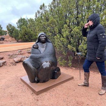 Allan Houser - A woman wearing a black puffer jacket, jeans, and brown boots stands outdoors on a dirt surface, talking on her phone. She is positioned to the right of a bronze sculpture depicting a woman with children, set against green bushes and a cloudy sky. The sculpture is mounted on a rectangular base, and the scene appears to be in a park or sculpture garden. - Art Galleries Near Me - Santa Fe, New Mexico