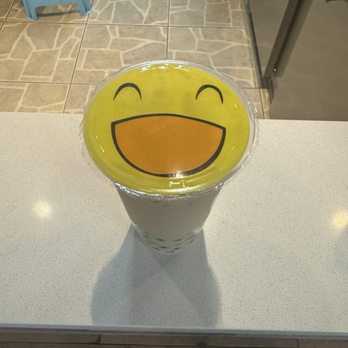 Tbaar - Bubble Milk Black Tea - A cup of bubble tea with black tapioca pearls at the bottom is positioned on a white countertop. The cup has a clear plastic lid featuring a large, yellow, smiling emoji face with closed eyes and an open mouth. The background shows tiled flooring, a person wearing sandals, and part of a stainless steel refrigerator or appliance. - Bubble Tea Near Me - Denver, Colorado