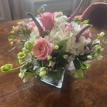 Mangel - A floral arrangement features pink roses, white hydrangeas, and additional small pink and green flowers with green leaves, all placed in a clear square glass vase. The bouquet is positioned on a polished wooden table, with a background showing a window with curtains and a radiator, indicating an indoor setting. - Florists Near Me - Chicago, Illinois