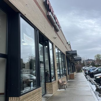Five Guys - The image shows the storefront of a Five Guys restaurant with large glass windows, a brick facade, and a sidewalk with benches and parked cars. - Burgers Near Me - Bayside, New York