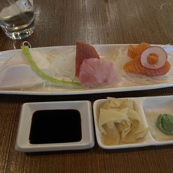 Yuzu Sushi - A17 - Mixed Sashimi (6 Pcs) - A rectangular ceramic plate contains slices of sashimi including tuna, salmon, and yellowtail, arranged on shredded daikon radish. Accompanying the sashimi are small dishes of soy sauce, wasabi, and pickled ginger. A glass of water is visible in the background on a wooden table, indicating a dining setting at a Japanese restaurant. - Japanese Near Me - Chamblee, Georgia