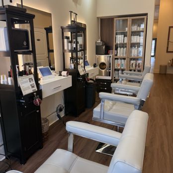 My Spa - The image shows a beauty salon interior with four white salon chairs arranged in a row, facing a counter with electronic devices and product displays. Behind the chairs, there are black and glass display cabinets containing hair and skincare products. The space has wooden flooring and neutral-colored walls, with additional shelves and equipment visible in the background. The setting appears clean and organized, suitable for hair or skincare treatments. - Day Spas Near Me - Chicago, Illinois