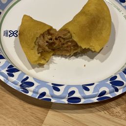 A meat-filled empanada, partially eaten, is placed on a decorative paper plate with blue and white patterns, on a wooden surface.