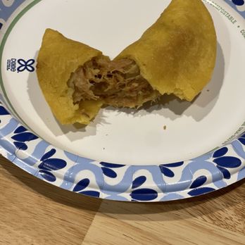 Sofia’s Empanadas - A meat-filled empanada, partially eaten, is placed on a decorative paper plate with blue and white patterns, on a wooden surface. - Colombian Near Me - East Meadow, New York
