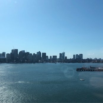 Hyatt Regency Boston Harbor - View from hotel - The image shows a panoramic view of Boston's downtown skyline across Boston Harbor, with numerous high-rise buildings and construction cranes. The water in the foreground reflects sunlight, and small boats are anchored near the docks. The scene is captured during daylight with a clear blue sky, indicating a bright, sunny day. - Hotels Near Me - Boston, Massachusetts