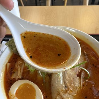 Yujo - Mmmmm... - A bowl of ramen featuring broth, a soft-boiled egg, sliced pork, and vegetables, with a spoon held above the bowl. - Ramen Near Me - Indianapolis, Indiana