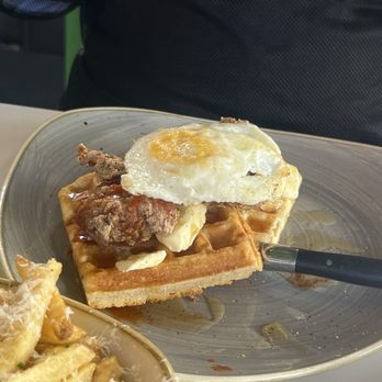 Everly - Chicken and Waffles - A waffle with fried chicken, a fried egg, and syrup is served on a gray plate, with a bowl of pasta nearby. - New American Near Me - Madison, Wisconsin