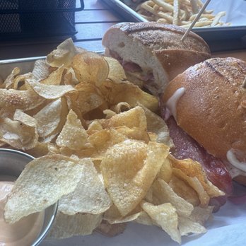 Commonwealth - Spicy Chicken sandwich and chips - A tray holds two sandwiches made with hoagie rolls, filled with meats and toppings, accompanied by a large portion of potato chips and a small metal container of dipping sauce. The setting appears to be a casual dining restaurant with a wooden table and additional food in the background. - Beer Bar Near Me - Modesto, California