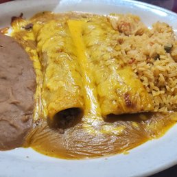 Enchilada - A white oval plate contains shredded beef enchiladas topped with melted cheese and sauce, alongside refried beans and Mexican rice with vegetables. The dish is served on a dark wooden table, with a red drink partially visible in the background.