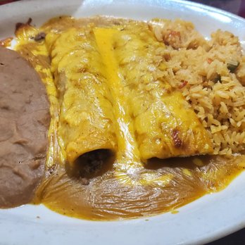 Los Generales - Shredded beef enchilada combo - A white oval plate contains shredded beef enchiladas topped with melted cheese and sauce, alongside refried beans and Mexican rice with vegetables. The dish is served on a dark wooden table, with a red drink partially visible in the background. - Mexican Near Me - Spring, Texas