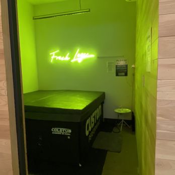 Custom Performance - The image shows a small, enclosed room with a neon sign on the wall that reads 'Fresh Leaf' in bright green. Below the sign is a cold storage unit labeled 'COLDTUB.' The room has a wooden floor, a small stool in the corner, and a sign on the wall indicating shower instructions. The lighting is primarily from the neon sign, creating a greenish glow. - Physical Therapy Near Me - New York, New York
