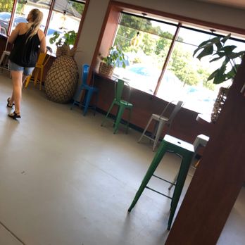 Botanas snack bar - Seating area - The image shows an indoor seating area with large windows allowing natural light, featuring a variety of colorful chairs in yellow, blue, green, and gray. Potted plants are placed on the window sills, and a woman with blonde hair, wearing a black top and denim shorts, is walking away from the camera. The space has a simple, modern decor with a neutral floor and ceiling tiles. - Ice Cream & Frozen Yogurt Near Me - Terrell, Texas