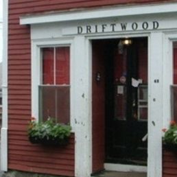the entrance to driftwood - The image depicts the exterior of Driftwood, a small business with red horizontal siding, white trim, and flower boxes on either side of the entrance.