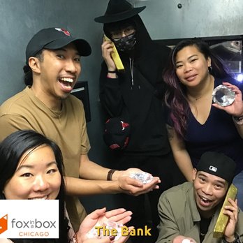 Fox in a Box - A group of five people is gathered inside an escape room, with a gray wall background. They are holding large, clear, crystal-like objects and phones, smiling and posing for the camera. The group includes both men and women, with casual clothing and accessories. The Fox in a Box Chicago logo is visible in the bottom left corner, indicating the location as an escape room venue specializing in themed private rooms such as Bunker, Bank, Prison, and Zodiac. - Escape Games Near Me - Chicago, Illinois