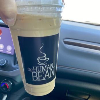 The Human Bean - Iced coffee - A plastic cup of iced coffee from The Human Bean is held inside a vehicle, with the car's dashboard visible in the background. - Coffee & Tea Near Me - Albuquerque, New Mexico