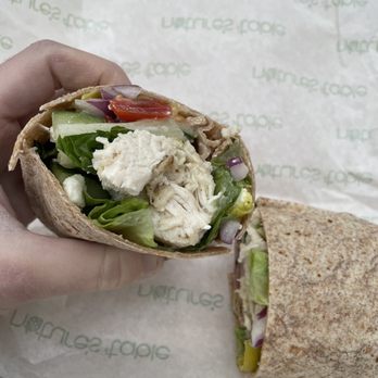 Nature's Table - Mediterranean Wrap - A chicken salad wrap filled with lettuce, tomato, cucumber, red onion, and chicken is held by a hand, with a second wrap on the paper. - Salad Near Me - Indianapolis, Indiana