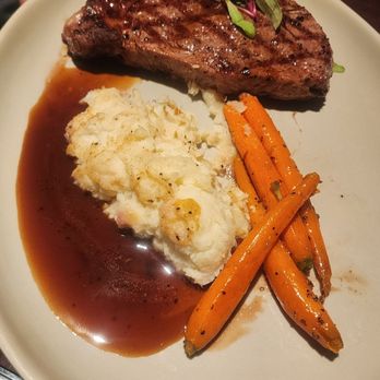 Red Sage - Ribeye - A dinner plate contains a grilled steak topped with microgreens, accompanied by mashed potatoes with gravy, and roasted carrots. The steak has grill marks, and the mashed potatoes are creamy with a brown gravy. The roasted carrots are seasoned and slightly charred. The dish is presented on a white plate, with a dark background visible at the top edge. - New American Near Me - Santa Fe, New Mexico