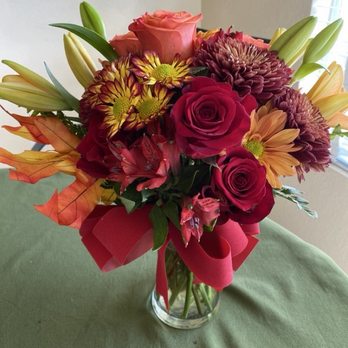 Arrowhead Flowers - A floral arrangement featuring roses, lilies, chrysanthemums, and other flowers in a glass vase, decorated with a red ribbon, placed on a green tablecloth. - Florists Near Me - Glendale, Arizona