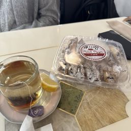 A glass cup of tea with a lemon wedge on a saucer, alongside a box of Polish paczki from Polish Paczki Cafe, on a table with decorative tiles.