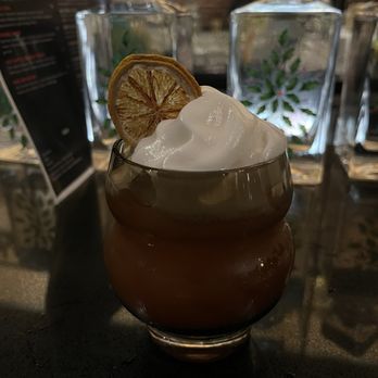 Fomo - A dark-colored cocktail in a rounded glass, garnished with a dried lemon slice and topped with whipped cream, set on a bar counter. - Cocktail Bars Near Me - Redding, California