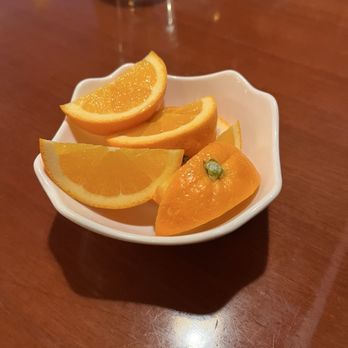 Nori - A small, white, scalloped-edge dish contains three orange slices and a yellow pepper, arranged on a polished wooden table. In the background, there is a clear glass of water and a dark object, possibly a menu or electronic device. The setting appears to be a restaurant or dining area. - Japanese Near Me - Montclair, New Jersey