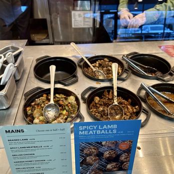 CAVA - A glass display case shows various cooked meats and vegetables, with a menu and promotional sign for spicy lamb meatballs in front. - Mediterranean Near Me - Cedar Park, Texas