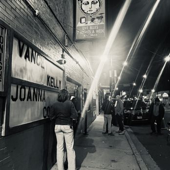 Kingston Mines - Night scene outside Kingston Mines, a historic blues nightclub in Chicago, with several people standing on the sidewalk. The image is in black and white, showing a sign for the club and a group of individuals near the entrance. Streetlights illuminate the scene, and parked cars are visible along the street. - Jazz & Blues Near Me - Chicago, Illinois