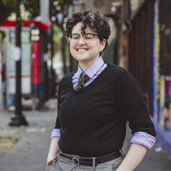 Wolff Osteopathic Medicine - Dr Wolff, a person with brown hair with a grey streak, black sweater, grey pants and purple collared shirt standing on a city street