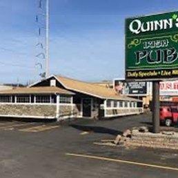 A hidden gem! A+ - The image depicts the exterior of Quinn's Irish Pub, featuring a large green sign with the pub's name, a building with a porch, and a parking lot with a few vehicles. The signage indicates it is a sports bar and Irish pub, with additional signs advertising daily specials and live music.