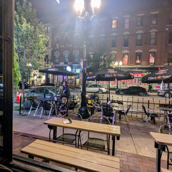 Lone Owl Pub - The image depicts an outdoor patio area with several tables and chairs, some with umbrellas branded Tito's Handmade Vodka, situated along a city sidewalk. A lamppost is centrally located, illuminating the scene. In the background, a busy street with parked and moving cars, storefronts, and multi-story buildings are visible. The scene is captured during nighttime, with artificial lighting and a few pedestrians present. - Cocktail Bars Near Me - Chicago, Illinois