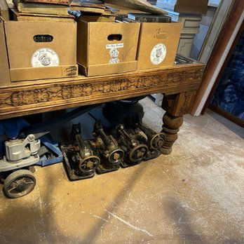 Lumber + Salt - A wooden table with ornate carved details holds three cardboard boxes labeled 'We Buy Books,' filled with books and topped with additional books and a piece of driftwood. The setting appears rustic, with a concrete floor showing signs of wear and vintage machinery placed underneath the table. In the background, there are cardboard boxes and a glimpse of a window with curtains, suggesting an antique or thrift shop environment. - Antiques Near Me - Riverhead, New York
